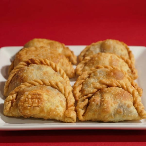 A white rectangular plate holds eight golden-brown, freshly baked Tango's Empanadas against a red background.